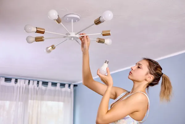 Vervanging van een LED-lamp in plafond kroonluchter thuis, jonge witte vrouw met nieuwe lamp in haar hand. — Stockfoto