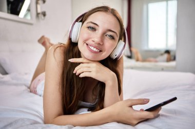 29 yaşında, kafasında kablosuz kulaklıkla gülümseyen bir kadın elinde telefonla yatağında yatıyor..