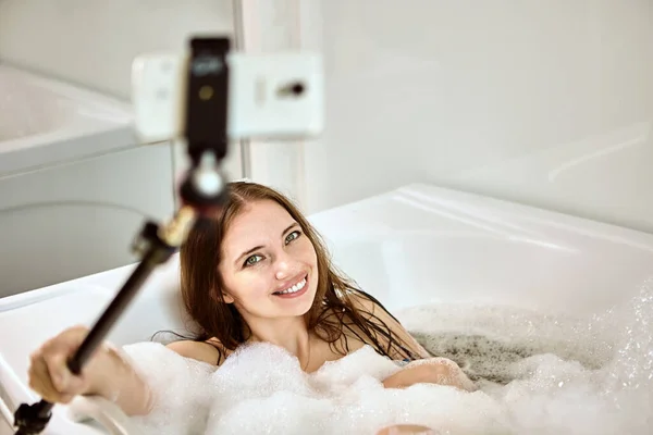 Bain tourbillon dans la chambre d'hôtel, femme communique en ligne en fixant son téléphone sur selfie stick. — Photo