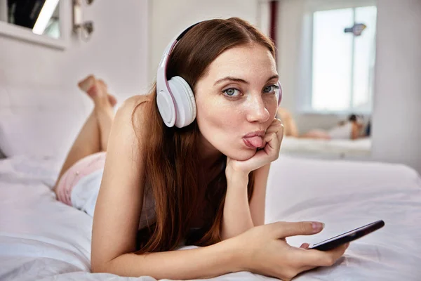Młoda kobieta wystaje z języka leżąc w łóżku w słuchawkach bezprzewodowych z telefonem w rękach. — Zdjęcie stockowe
