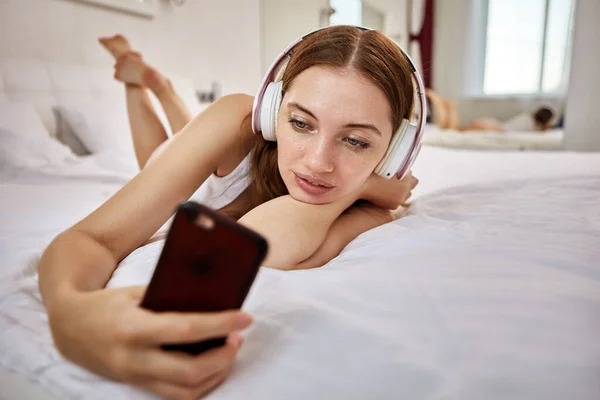 Kobieta ze smartfonem i bezprzewodowymi słuchawkami leży w łóżku na brzuchu i wykonuje rozmowy wideo. — Zdjęcie stockowe