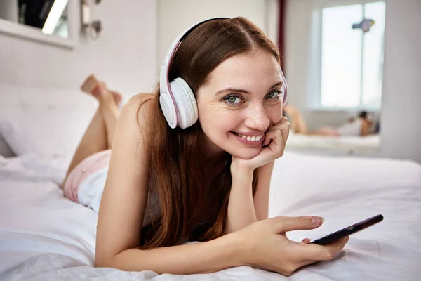 Glimlachende vrouw met draadloze koptelefoon en smartphone ligt in bed op haar buik. — Stockfoto