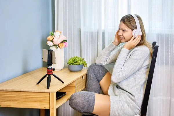 坐在椅子上笑着的无线耳机中的女人在三脚架上方便地使用视频通话 图库图片