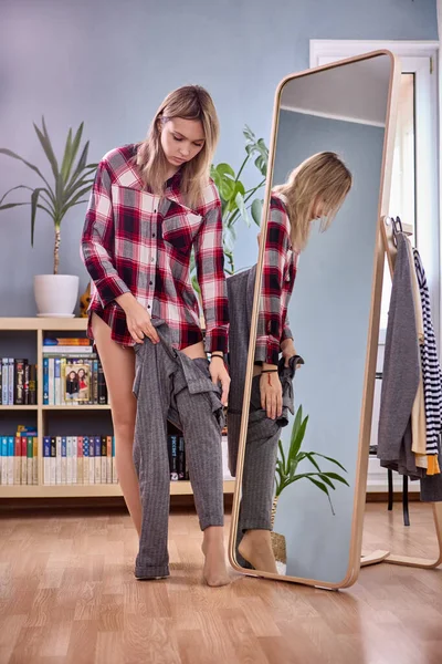 Fitting in front of full length mirror by slim woman in checkered shirt in bedroom at home.