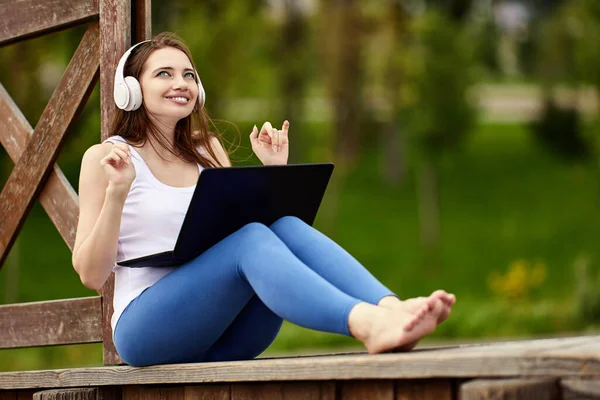 Online möte i naturen av glada kvinna i sladdlösa hörlurar med bärbar dator utanför. — Stockfoto