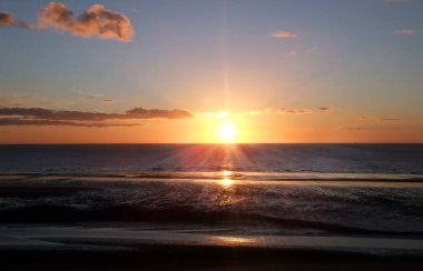 Işıl ışıl parlayan bir günbatımı, sakin mavi bir deniz ve plajın üzerinde ışıl ışıl parlayan turuncu bulutlarla