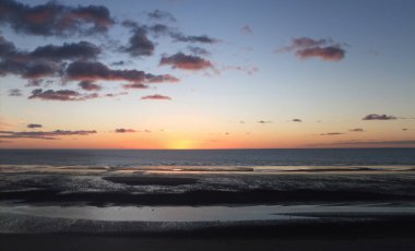 Geniş panoramik alacakaranlık sahnesi. Koyu pembe günbatımı gökyüzü ve bulutlar sahilde sakin bir deniz ve suya yansıyor.