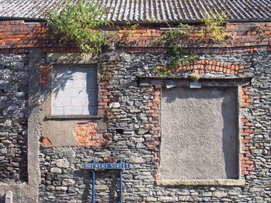 Kapalı camları olan terk edilmiş eski bir taş bina çatıda yabani otlar büyüyor ve Ulverston 'daki bira fabrikası sokağında bir tabela var.