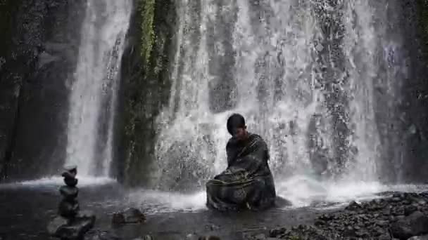 一个亚洲人坐在山河的岩石上沉思的画像 在一个新鲜的瀑布下 放松地清理你的思绪 享受凉爽的自然美 — 图库视频影像