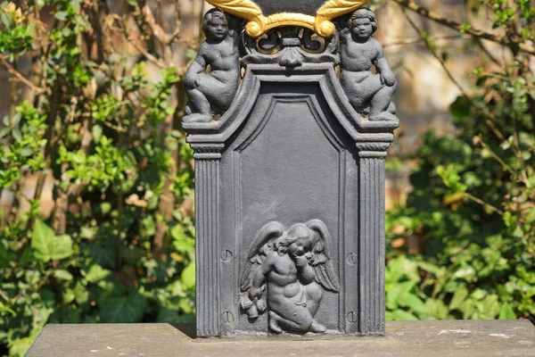 Base of a grave cross from the Art Nouveau period. Two wingless putti kneel above the stylized columns and look at the viewer. At the foot, a little angel kneels behind his back, holding some poppy seed pods.