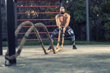 Genç yakışıklı bir adam savaş halatıyla çalışıyor. Sağ kavrayış ve sağlam duruş gösteriyor. Egzersiz maskesi takıyor, dışarıda egzersiz yapıyor.