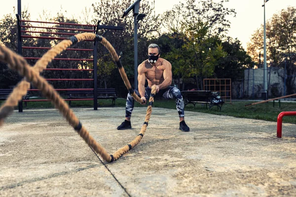 Genç kaslı kaslı adam savaş halatı egzersiziyle dayanıklılığını geliştiriyor. Solunum direnci maskesi takıyor. Açık hava sporu konsepti.