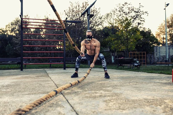 Genç kaslı kaslı adam savaş halatı egzersiziyle dayanıklılığını geliştiriyor. Solunum direnci maskesi takıyor. Açık hava sporu konsepti.