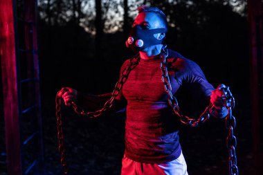 Güzel görünüşlü, güçlü, halter zincirleriyle parkta çalışan, yüksek irtifa simülasyon maskesi takan, dayanıklı ve dayanıklı bir adam.