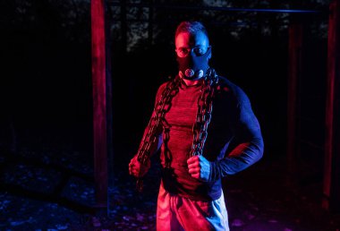 Yakışıklı kaslı bir adam solunum maskesi takmış ve halter zincirlerini vücuduna sarmış halde gece sporu konseptini tutarken korkunç bir bakış atıyor.