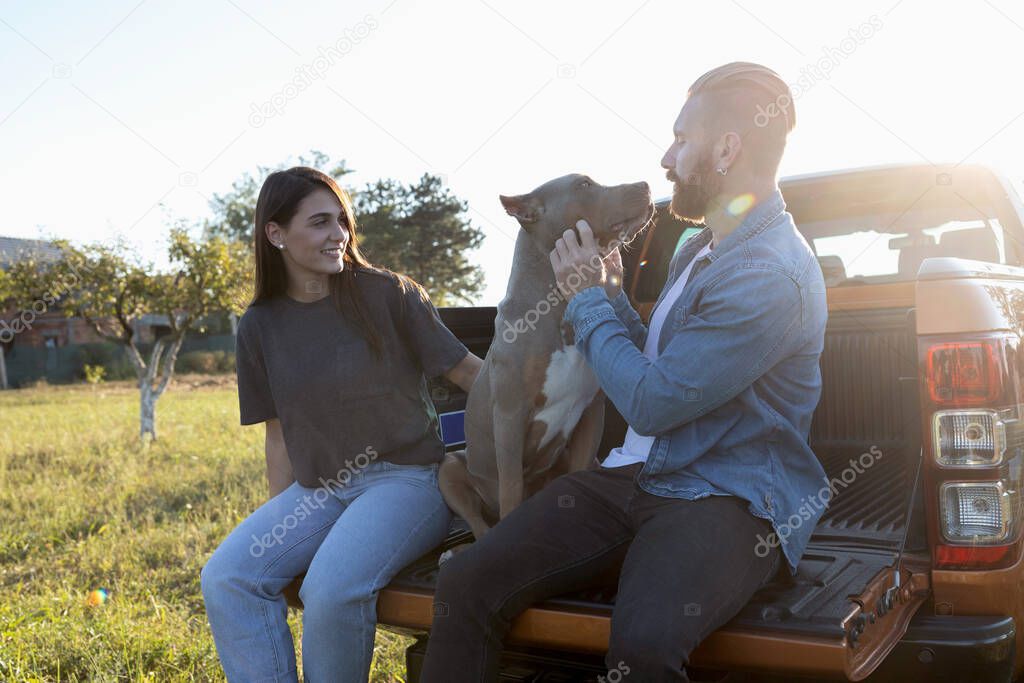 Cute young couple and their canine bully dog spending time together while sitting on the back of a pickup truck with sun shining in their backs