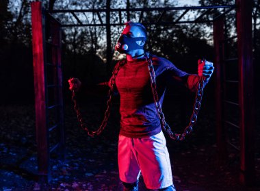 Parkta alacakaranlıkta halter zincirleriyle çalışan yakışıklı, korkutucu kaslı adam. Maske takıyor, spor yapıyor, park konseptinde sokak çalışması yapıyor.