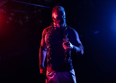 Yakışıklı kaslı bir adam solunum maskesi takmış ve halter zincirlerini vücuduna sarmış halde gece sporu konseptini tutarken korkunç bir bakış atıyor.