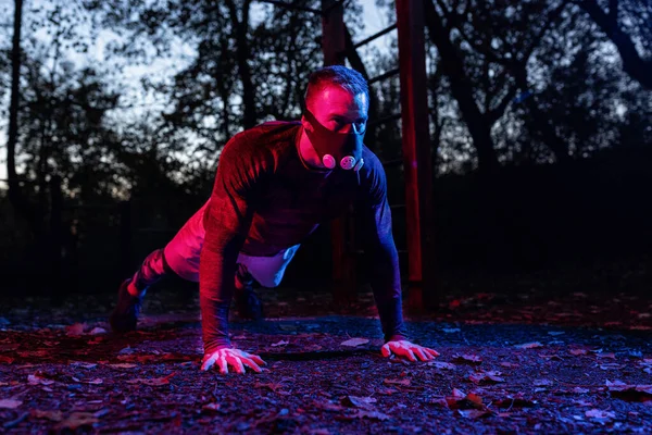 Yüksek rakımlı simülasyon maskesi takan deneyimli bir atlet. Akşam karanlığında parkta şınav çekiyor. Fonksiyonel egzersiz, sokak göğsü egzersizi.