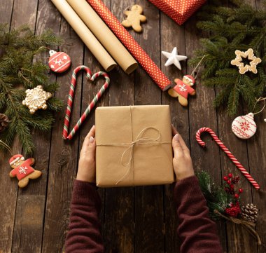 Elinde Noel hediyesini, şeker kamışı, zencefilli kek bayramı erkeği, Noel kurabiyeleri ve diğer çeşitli festival şekilli bisküviler, hediye verme ve paylaşma konsepti gibi kış nedenleriyle süslenmiş ahşap masanın üzerinde tutan kadın.