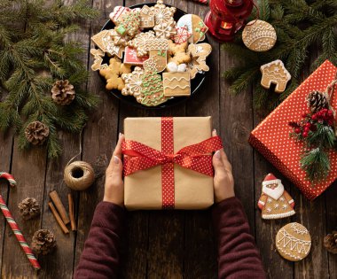 Güzel dekore edilmiş bir Noel hediyesini köylü ahşap masasında, kış tatili motifleriyle şekillendirilmiş şeker şenlikli zencefilli kurabiyelerle dolu bir tabağın yanında tutan kadın, keyifli tatil kavramı.