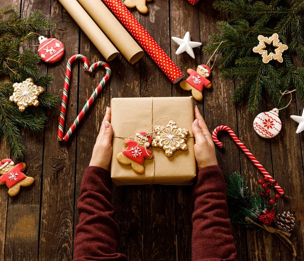 Kış tatili motifleri, bisküviler, şekerler ve diğer süslemelerle süslenmiş ahşap bir masanın üzerinde Noel hediyesini tutan kadınların ellerini kapatın.