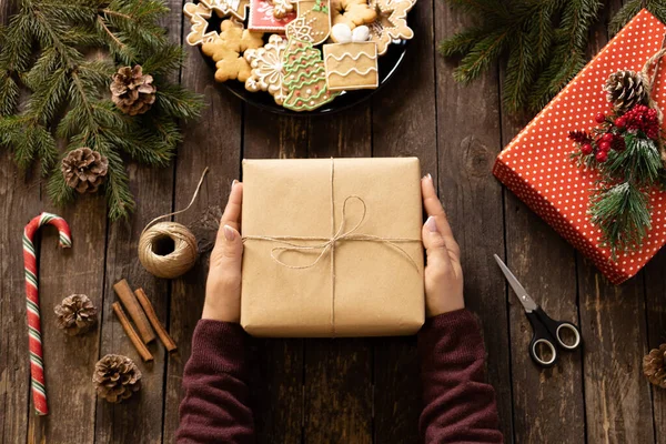 Bir kadının elleri, elinde eko kağıda sarılı bir hediye kutusuyla kırsal ahşap masa üzerinde kış kurabiyeleri ve süslemeleri olan, süslü bir konsept taşıyor.