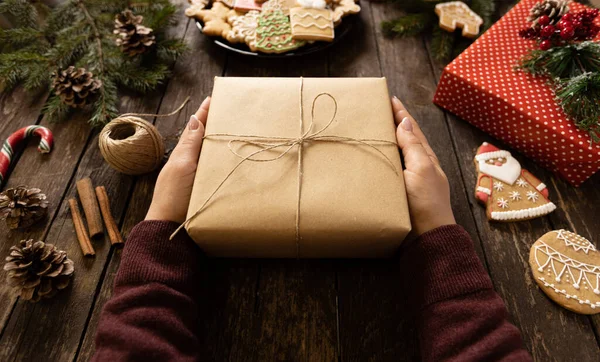 Bir kadının elleri, elinde eko kağıda sarılı bir hediye kutusuyla kırsal ahşap masa üzerinde kış kurabiyeleri ve süslemeleri olan, süslü bir konsept taşıyor.