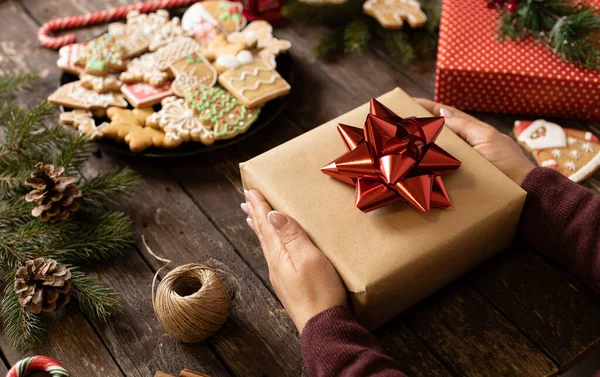 Kadın eli, Noel hediyesini çeşitli bayram şekillerinde ev yapımı lezzetli kurabiyelerin yanında tutuyor. Ahşap arka plan kavramı.