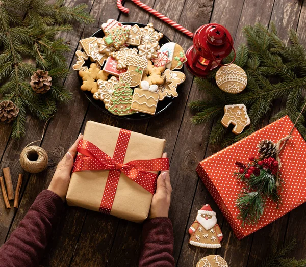 Noel hediyesini tutan eller güzel dekore edilmiş ve ahşap bir masaya sarılmış şenlikli kış kurabiyeleri ve bisküvilerle dolu bir tabağın yanında, Noel temalı konsept.