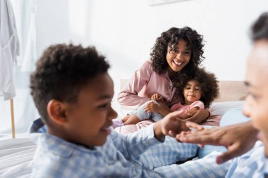 Neşeli Afro-Amerikalı kadın kızı yatakta kucaklıyor baba oğul bulanık ön planda eğleniyorlar.