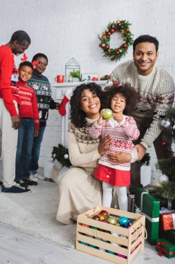 Afro-Amerikan kızı, Noel balosunu mücevherlerle dolu ahşap bir kutunun yanında tutuyor ve oturma odasında mutlu bir aile var.