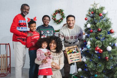 Noel ağacı ve Afrika kökenli Amerikan ailesinin yanında süslü mücevherlerle dolu tahta kutu tutan adam.