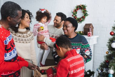 Çocuk, oturma odasında, neşeli Afro-Amerikan ailesinin yanında içinde noel topları olan tahta bir kutu tutuyor.