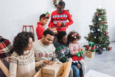 Neşeli Afro-Amerikan ailesi oturma odasında bulanık Noel ağacıyla hediyeler paketliyor.