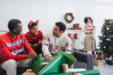 Ren geyiği boynuzları takmış afro amerikalı çocuk hediye paketleyen adamların yanında gülümsüyor ve noel ağacının yanında annesini bulanıklaştırıyor.