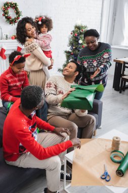 Gülümseyen Afrikalı Amerikalı kadın Noel hediyesi paketleyen adamın elini gösteriyor.