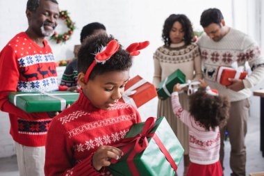 Ren geyiği boynuzları takmış afrikalı Amerikalı çocuk elinde hediye kutularıyla bulanık bir ailenin yanında Xmas tutuyor.