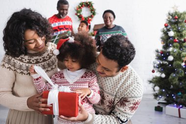 Noel Baba şapkalı Afro-Amerikalı kız mutlu ebeveynlerin yanında hediyesini tutuyor ve oturma odasında Noel ağacıyla ailesini bulanıklaştırıyor.