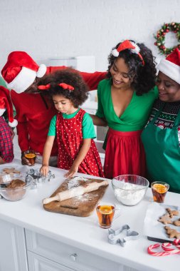 Noel Baba şapkalı mutlu Afro-Amerikan ailesi Noel kurabiyeleri hazırlayan kızın yanında.