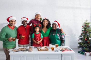 Noel Baba şapkalı neşeli Afro-Amerikan ailesi mutfakta Noel kurabiyeleri hazırlarken kameraya bakıyor.