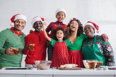 Afrikalı Amerikalı kız Noel Baba şapkası ve malzemeleriyle mutlu bir ailenin yanında şarkı söylüyor.