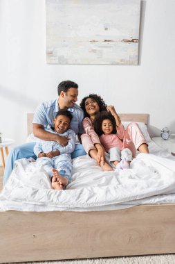 Mutlu Afro-Amerikalı çift pijamalarıyla çocukları kucaklıyor ve yatak odasında gülüyor.