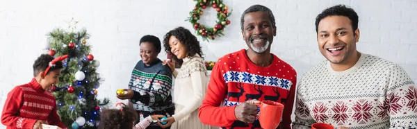 快乐的非洲男人 拿着杯子站在模糊的家庭旁装饰着圣诞树和横幅 — 图库照片