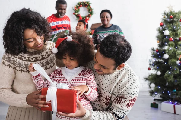 Noel Baba şapkalı Afro-Amerikalı kız mutlu ebeveynlerin yanında hediyesini tutuyor ve oturma odasında Noel ağacıyla ailesini bulanıklaştırıyor.