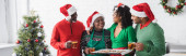 středního věku africký Američan žena drží pečení list s vánočními cookies v blízkosti šťastné rodiny a vánoční strom, banner