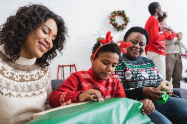 Ren geyiği boynuzları takan Afrikalı Amerikalı çocuk mutlu anne ve büyükannenin yanında noel hediyesi paketliyor.