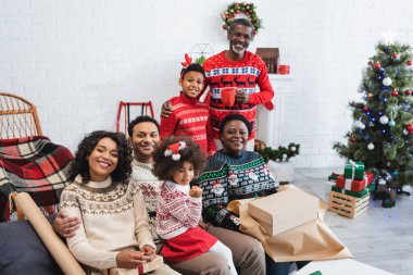Neşeli Afro-Amerikan ailesi, bulanık noel ağacının yanındaki hediye kutularını paketlerken kameraya bakıyor.