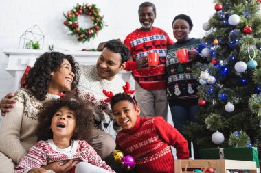 Afro-Amerikan ailesine bakıp Noel ağacının yanında gülen fincanları olan mutlu büyükanne ve büyükbaba.