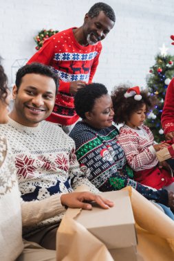 Orta yaşlı bir adam Afrika kökenli Amerikan ailesine bakıyor. Noel hediyeleri paketliyor.